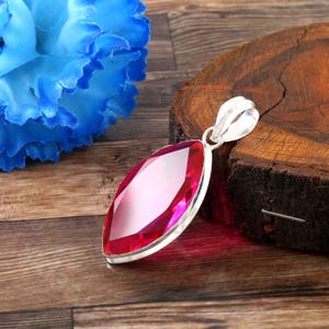 Pode incluir: Um pingente com uma gema rosa vibrante em lapidação marquise, engastada numa moldura de prata polida com uma argola resistente no topo. A pedra facetada reflete a luz, exibindo um tom magenta profundo e uma forma alongada com pontas.