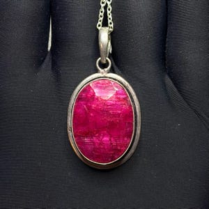 May include: An oval-shaped pendant featuring a faceted, deep magenta-red gemstone set in a polished silver-toned metal frame. The pendant hangs from a delicate silver-toned chain, showcasing a smooth, reflective surface and a vibrant, saturated colour.