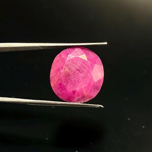 Könnte beinhalten: Ein leuchtender, oval geschliffener Edelstein in einem tiefen Magentarosa mit facettierter Oberfläche. Der Stein wird horizontal von einer silberfarbenen Metallpinzette gehalten und zeigt deutlich seine Transparenz und Struktur.