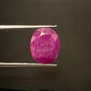 Könnte beinhalten: Ein oval geschliffener, tief magentafarbener Edelstein mit facettierter Oberfläche, der horizontal von einer silberfarbenen Metallpinzette gehalten wird. Der Stein weist sichtbare interne Einschlüsse und eine polierte Oberfläche auf, die das Licht reflektiert.