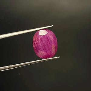 Op de afbeelding: Een levendige, ovaalvormige, gefacetteerde robijn met een diepe magenta-rode tint. De edelsteen wordt stevig vastgehouden tussen de twee dunne, zilverkleurige metalen punten van een pincet, wat de doorschijnende kwaliteit en het gepolijste oppervlak toont.