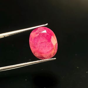 May include: An oval-shaped, faceted gemstone with a vibrant, deep pinkish-red hue. The stone displays a complex internal structure with visible natural inclusions and light-reflecting facets across its surface, held by metal tweezers.