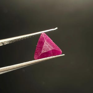 Può includere: Una gemma di un vibrante rosso rosato intenso, tagliata in forma triangolare. La pietra presenta molteplici superfici sfaccettate che catturano la luce, mostrando un colore ricco e saturo in tutta la sua struttura. Tenuta da pinzette metalliche.