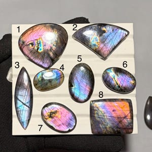 Könnte beinhalten: Acht polierte Labradorit-Edelsteine in verschiedenen Formen und Größen, angeordnet auf einem weißen, gerillten Display. Jeder Stein zeigt irisierende Blitze in Violett, Blau und Gold, mit nummerierten Etiketten von eins bis acht.