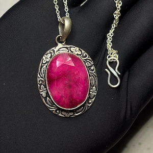 May include: A necklace featuring an oval-shaped, faceted, deep pink gemstone pendant set in a dark, oxidised silver-toned metal frame. The frame is decorated with a raised floral and leaf pattern. The pendant hangs from a thin silver-toned chain.