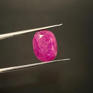 Pode incluir: Uma gema de rubi de corte oval com um tom vermelho rosado profundo e superfície multifacetada. A gema é mantida firmemente entre as pontas de uma pinça de metal prateado, exibindo sua qualidade translúcida e inclusões internas visíveis.