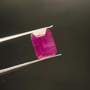 Könnte beinhalten: Ein rechteckig geschliffener, leuchtend magentafarbener Edelstein, der von einer silberfarbenen Metallpinzette gehalten wird. Der Stein hat eine facettierte Oberfläche mit sichtbaren Einschlüssen und ein poliertes Finish in satter Farbe.