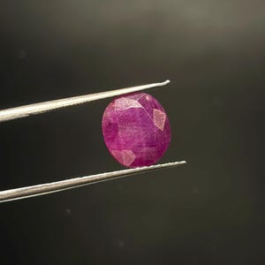 Könnte beinhalten: Ein facettierter, oval geformter, tief pink-roter Edelstein, der von einer silberfarbenen Metallpinzette gehalten wird. Der Stein zeigt eine durchscheinende Qualität mit sichtbaren Einschlüssen und eine polierte Oberfläche, die Licht reflektiert.