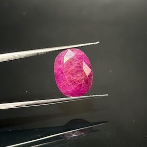 Puede incluir: Una gema de rubí de forma ovalada con una superficie facetada, que muestra un tono rojo rosáceo intenso e inclusiones internas visibles en forma de aguja, sostenida entre las puntas de unas pinzas de metal plateado.