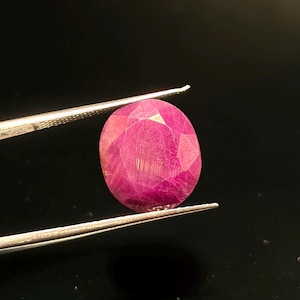 Op de afbeelding: Een gefacetteerde, ovaalvormige robijn met een diepe roze-rode tint. De steen vertoont een complex patroon van geometrische slijpvlakken over het oppervlak, die het licht reflecteren en de interne structuur en levendige kleur benadrukken.