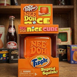 K&ouml;nnte beinhalten: Ein Dr. Pepper Nee Doh Nice Cube Spielzeug in orangefarbener Verpackung. Die Schachtel zeigt den Text 'Nee Doh', 'Nice Cube' und 'Peppte Suptid Squish'. Das Spielzeug ist ein glattes, orangefarbenes, quadratisches Quetschobjekt hinter einem Sichtfenster.