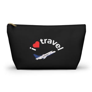 Può includere: Pochette da viaggio nera con cerniera dorata. La pochette presenta una grafica bianca con le parole "I love travel", un cuore rosso e un aereo bianco e blu.