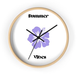 Può includere: Orologio da parete rotondo con cornice in legno chiaro. Il quadrante bianco presenta un fiore di ibisco viola chiaro e le parole "Summer" e "Vibes" in nero, con lancette nere.