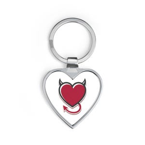 Può includere: Un portachiavi in metallo a forma di cuore con sfondo bianco e un grafico a cuore rosso al centro. Il design del cuore include piccole corna da diavolo nere sulla parte superiore e una coda appuntita sul fondo, in una cornice color argento.