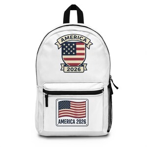 Può includere: Zaino bianco con manico e cerniera neri. La parte anteriore presenta un disegno a scudo con la bandiera americana e la scritta "AMERICA 2026". È presente anche una toppa rettangolare più piccola con la bandiera americana e la scritta "AMERICA 2026".