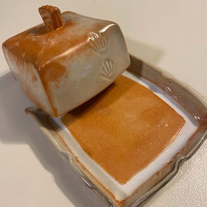 May include: A ceramic butter dish featuring a rectangular base and a matching lid with a small handle. The piece has a white glaze with mottled orange and rust-colored accents, and the lid is decorated with small, stamped shell-like impressions.