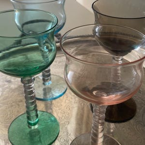 Puede incluir: Un conjunto de cuatro copas con tallos acanalados y bases circulares. Las copas presentan cuencos translúcidos en colores distintos, incluyendo verde, rosa, transparente y gris humo oscuro, cada una con un borde liso y forma redondeada.