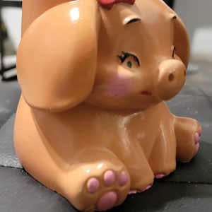 May include: A peach-colored ceramic planter shaped like a sitting piglet. The piglet features a small red bow on its head, painted pink cheeks, and pink paw pads on its feet. The surface has a smooth, glossy finish with a hollow opening at the top.