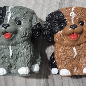 May include: Two small dog figurines with curly textured fur. One dog is grey with black ears and a white stripe on its forehead and chest. The other dog is brown with black ears and a white stripe on its forehead and chest. Both have white paws.
