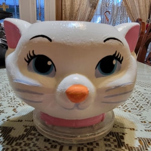 May include: A white ceramic planter shaped like a cat head with large blue eyes, long black eyelashes, and a bright orange nose. The cat features pink inner ears and a pink collar around its neck, resting on a clear circular base for stability.