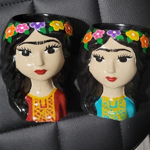 May include: Two ceramic head planters featuring dark hair adorned with a colorful floral crown. One figure wears a red top with yellow geometric patterns, while the other wears a blue top with yellow patterns. Both have painted facial features.