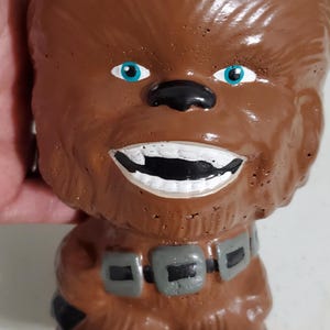 May include: A brown ceramic mug shaped like the head and torso of the Star Wars character Chewbacca. The mug features textured fur details, bright blue eyes, a wide white-toothed grin, and a grey utility belt with black square accents.