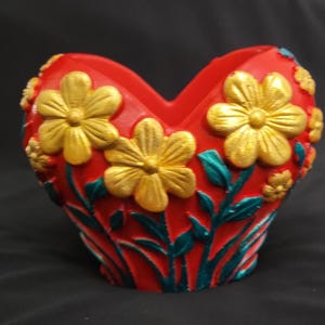 May include: A heart-shaped red vase featuring raised gold-colored flowers and teal-colored stems and leaves. The vessel has a wide, open top with a central dip, and the floral design wraps around the exterior surface of the red container.