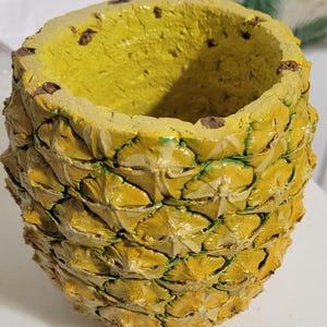 May include: A cylindrical planter shaped like a pineapple with a textured, yellow and green exterior. The interior is hollow and painted a solid yellow. The surface features a repeating geometric pattern of scales, mimicking the appearance of a pineapple.