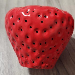 May include: A bright red ceramic planter shaped like a strawberry, featuring a textured surface with numerous small indentations and painted black dots scattered across the exterior to mimic the appearance of strawberry seeds.