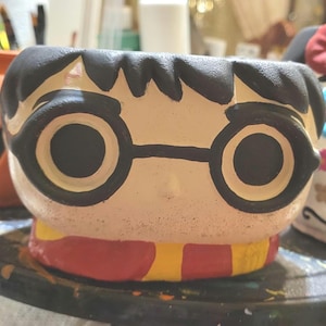 May include: A ceramic planter shaped like a head with black hair, large round black-rimmed glasses, and a red and yellow striped scarf at the base. The face is painted in a light beige tone with black details for the hair and glasses.
