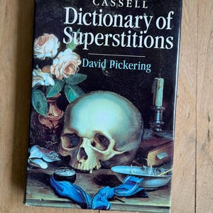 May include: A hardback book titled Cassell Dictionary of Superstitions by David Pickering. The black cover features a still life illustration of a human skull, a vase with white roses, a lit candle, a small bowl, and a bright blue ribbon.
