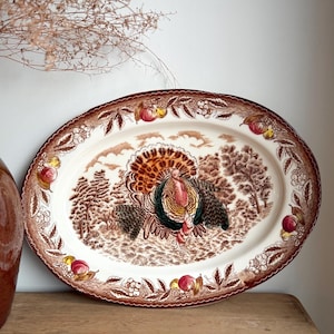 May include: An oval ceramic serving platter featuring a central illustration of a turkey in a landscape. The rim is decorated with a brown transferware pattern of fruits, leaves, and berries, accented with touches of red and yellow colour.
