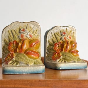 May include: A pair of heavy, arched bookends featuring a raised, three-dimensional floral relief. The design displays orange and yellow tulips with green leaves against a textured, speckled background, finished with a blue base at the bottom.