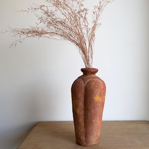 May include: A tall, terracotta-coloured vase with a textured, weathered surface and vertical fluted panels. The vase features patches of yellow discolouration and holds a large, airy arrangement of thin, dried brown branches.