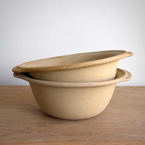 Peut inclure: Deux bols en céramique beige empilés avec une finition mate. Chaque bol présente un large bord arrondi et de petites poignées latérales intégrées. Les bols ont une silhouette incurvée simple et une couleur beige uniforme sur toute la surface.