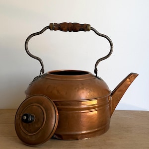 May include: A copper kettle with a rounded body, a short spout, and a removable lid featuring a small black knob. The kettle has a curved metal handle with a central wooden grip. The surface of the copper shows a warm, metallic finish.