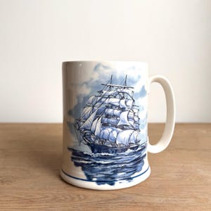 Peut inclure: Une tasse en céramique blanche ornée d'une illustration détaillée en bleu et blanc représentant un grand voilier à plusieurs mâts et voiles déployées sur l'océan. Le motif inclut des nuages bleus et une ligne bleue à la base.