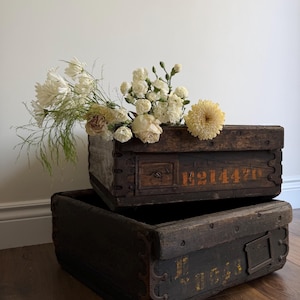 May include: Two aged wooden boxes, one on top of the other, with metal corner details and faded orange text. The top box is filled with a bouquet of white and yellow flowers, creating a rustic and charming display.