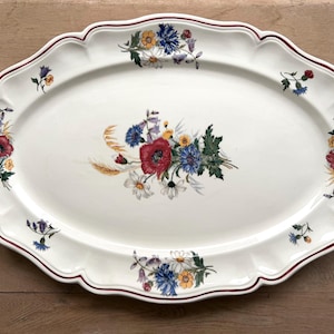 May include: An oval-shaped white ceramic serving platter with a scalloped edge and a thin dark red rim. The centre and the border feature colourful floral illustrations of red poppies, blue cornflowers, white daisies, and green foliage.