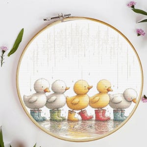 May include: Cross-stitch embroidery of five ducklings in colorful rain boots, standing in the rain. The ducklings are white and yellow, with orange beaks. The embroidery is set in a wooden hoop, with a white background.