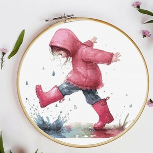 Op de afbeelding: Kruissteekborduurwerk van een kind in een roze jas met capuchon en regenlaarzen dat in een plas springt. Het kunstwerk is ingelijst in een houten borduurring, met spatten water en regendruppels. Het kleurenpalet omvat roze, blauw en wit.