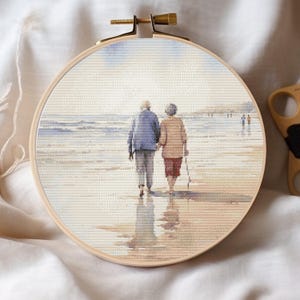 May include: A cross-stitch embroidery hoop depicting a tranquil beach scene. Two figures, hand-in-hand, stroll along the shore. The artwork is framed in a wooden hoop with a brass clasp. The colour palette includes soft blues, beige, and muted tones.