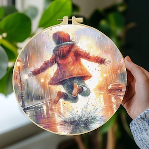 Op de afbeelding: Een ronde borduurring met een kleurrijk kruissteekpatroon van een persoon in een feloranje jas die tijdens een regenbui in een plas springt. De afbeelding bevat lantaarnpalen, een auto en een levendige, textuurrijke artistieke stijl.
