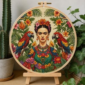 May include: A circular embroidery hoop containing a cross-stitch portrait of a woman with a floral crown, flanked by two red, blue, and yellow parrots. The design features vibrant green foliage and colorful flowers on a cream-colored fabric background.