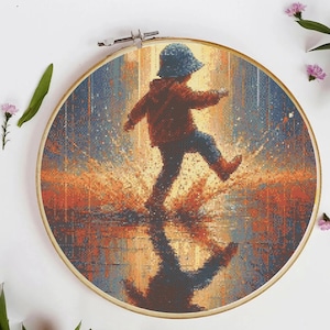 Op de afbeelding: Een borduurring met kruissteek met een kind in een rode jas en een blauwe hoed, halverwege een stap, spetterend in water. Het ontwerp gebruikt tinten oranje, blauw en bruin, met de weerspiegeling van het kind zichtbaar. De ring is van hout.