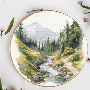 May include: A circular wooden embroidery hoop containing a cross-stitch pattern of a mountain landscape. The design features a flowing stream, rocky banks, a dirt path, and dense evergreen trees leading towards a large, pale purple mountain peak.