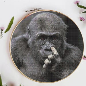 May include: A monochrome cross-stitch of a gorilla, with its finger touching its nose. The artwork is presented in a round wooden embroidery hoop, set against a white backdrop with pink flowers.
