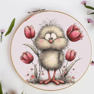 Peut inclure: Broderie au point de croix dans un cercle en bois représentant un poussin de dessin animé aux joues roses et aux pieds rouges, entouré de tulipes roses et de petites fleurs. Le fond est en tissu rose clair, pour un thème printanier fantaisiste.