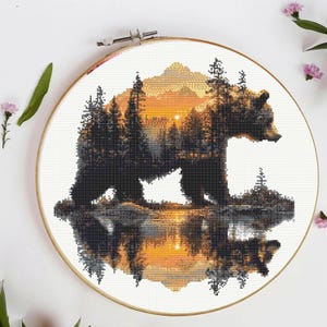 May include: A circular wooden embroidery hoop containing a cross-stitch pattern of a bear silhouette. The bear is filled with a landscape of dark pine trees and golden mountains against a sunset sky, with a reflection visible in the water below.