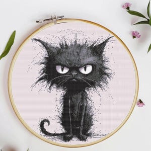 Op de afbeelding: Kruissteekborduurwerk van een chagrijnige zwarte kat met witte ogen, gezet in een houten hoepel. De kat heeft een strenge uitdrukking en stekelig haar. De achtergrond is een lichtroze stof.